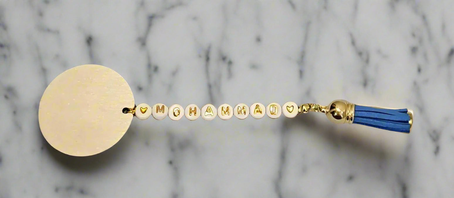 A customised wooden mohr with a blue tassel and the name 'Mohammad' written on it, with a round wooden disc attached to a metal clip.